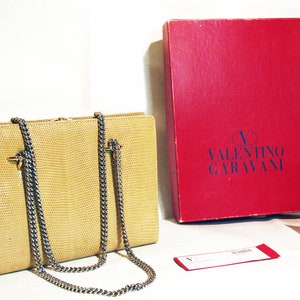 Puede incluir: Un bolso de mano de piel de serpiente beige con una correa de cadena plateada y herrajes dorados. El bolso est&aacute; sobre una superficie blanca junto a una caja roja con el texto "VALENTINO GARAVANI" en azul. Tambi&eacute;n hay una caja roja m&aacute;s peque&ntilde;a con el mismo texto.