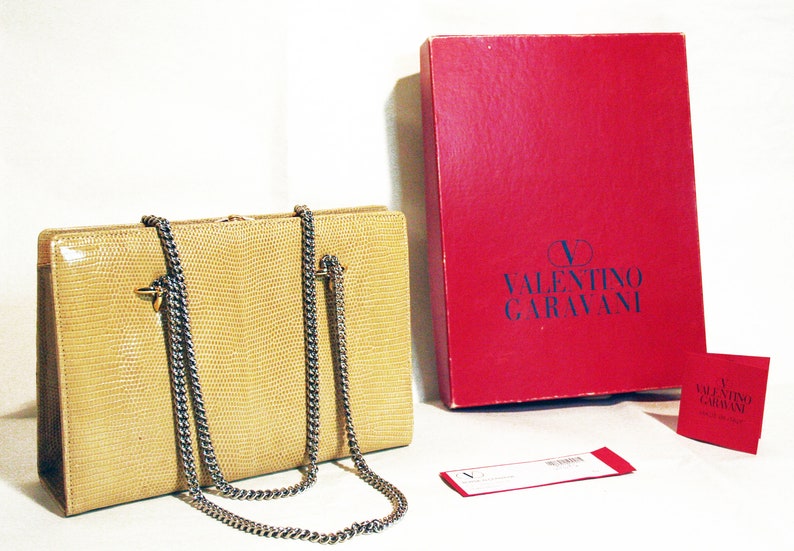 Puede incluir: Un bolso de mano de piel de serpiente beige con una correa de cadena plateada y herrajes dorados. El bolso est&aacute; sobre una superficie blanca junto a una caja roja con el texto "VALENTINO GARAVANI" en azul. Tambi&eacute;n hay una caja roja m&aacute;s peque&ntilde;a con el mismo texto.