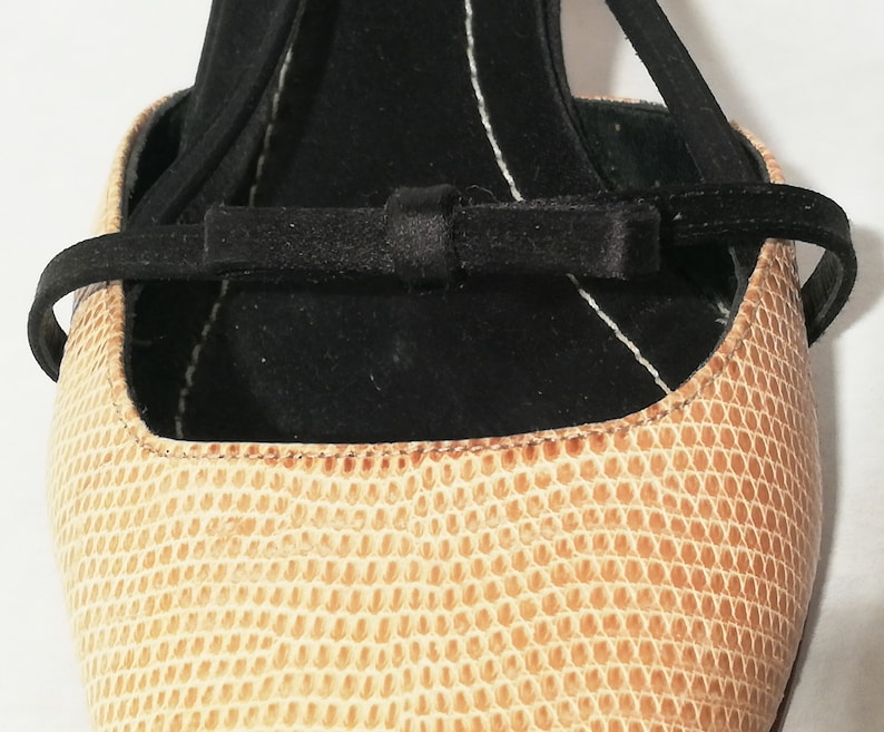 Puede incluir: Primer plano de un zapato beige texturizado con una correa negra y un detalle de lazo. El zapato tiene un patr&oacute;n similar al de un reptil y un interior negro. La correa se envuelve alrededor del tobillo.