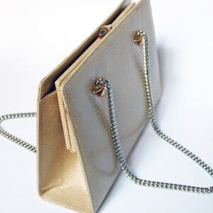 Puede incluir: Un bolso de mano beige de piel de serpiente con una correa de cadena plateada. El bolso tiene un cierre de broche y una forma rectangular.