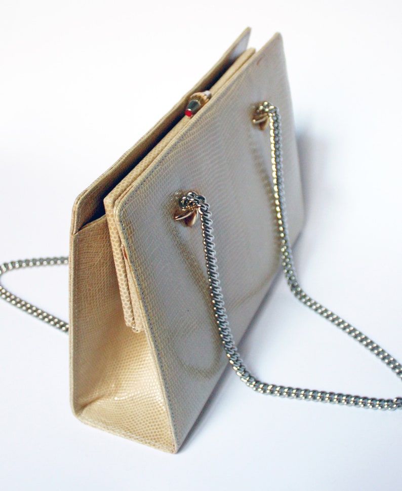 Puede incluir: Un bolso de mano beige de piel de serpiente con una correa de cadena plateada. El bolso tiene un cierre de broche y una forma rectangular.