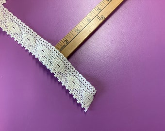 May include: Ivory-colored lace trim with a scalloped edge and rectangular cutouts. A wooden ruler with inch markings is positioned diagonally across the image. The background is a solid purple color. This trim is suitable for sewing and craft projects.