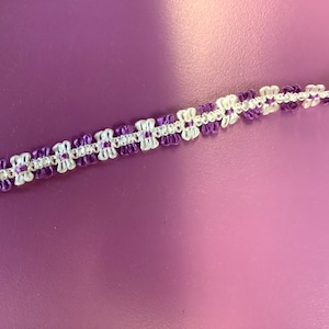 May include: A white and purple trim with a repeating flower pattern. The trim is made of a soft, woven fabric.