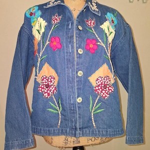 May include: A blue denim jacket featuring colorful fabric floral appliqués, embroidered stems, and decorative buttons. The front displays various flower shapes in pink, red, and blue patterns, with light-colored stitching along the collar.