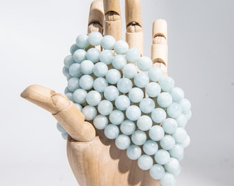 Pode incluir: Um colar ou pulseira de contas de água-marinha azul claro, redondas e lisas. As contas estão dispostas em um padrão simétrico.