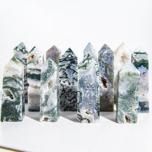 Puede incluir: Un grupo de 11 puntas de cristal de ágata verde y blanco dispuestas en una fila sobre una superficie blanca. Las puntas son de diferentes tamaños y formas, pero todas están pulidas y tienen un acabado liso y brillante.