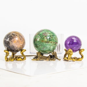 May include: Three decorative spheres on ornate gold metal stands. The spheres are made of different types of stone: a green and brown sphere, a purple sphere, and a gray sphere.