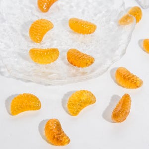Puede incluir: Una colecci&oacute;n de peque&ntilde;os segmentos de color naranja transl&uacute;cido con forma de gajos de c&iacute;tricos. Estas piezas decorativas tienen una superficie texturizada y realista, y est&aacute;n esparcidas tanto sobre un plato de vidrio transparente como sobre una superficie blanca.