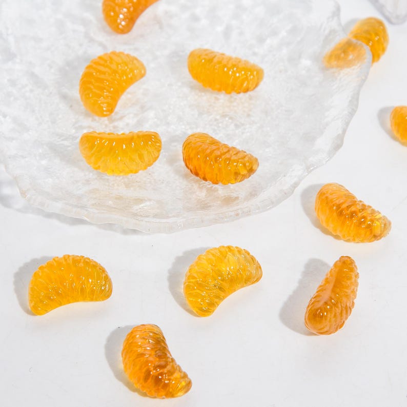 Puede incluir: Una colecci&oacute;n de peque&ntilde;os segmentos de color naranja transl&uacute;cido con forma de gajos de c&iacute;tricos. Estas piezas decorativas tienen una superficie texturizada y realista, y est&aacute;n esparcidas tanto sobre un plato de vidrio transparente como sobre una superficie blanca.