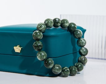 May include: A beaded bracelet featuring dark green, spherical beads. The beads have a marbled appearance with lighter green and white veining. The bracelet is displayed on a teal jewelry box with a gold crown emblem.