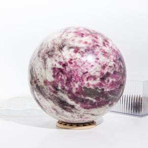 Puede incluir: Una gran esfera pulida de lepidolita rosa y blanca con patrones en espiral. La esfera está sobre un soporte de madera.