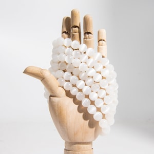 May include: A white gemstone bead necklace with round beads, displayed on a wooden hand.