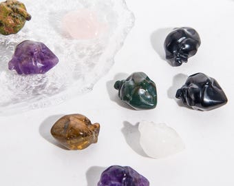 Op de afbeelding: Assortiment van gebeeldhouwde edelsteenbeeldjes in verschillende kleuren, waaronder amethist, rozenkwarts en groen. De beeldjes worden getoond op een wit oppervlak en een heldere glazen schaal. De edelstenen hebben een gladde, gepolijste afwerking. Afmetingen in centimeters.