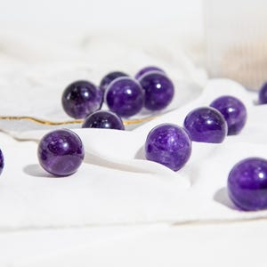 May include: A collection of amethyst crystal spheres in various sizes, displayed on a white fabric surface. The spheres are a deep purple colour and have a smooth, polished finish.