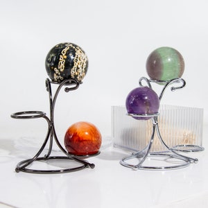 May include: Three polished gemstone spheres on metal stands. The spheres are black and white, green, and purple. The stands are black and silver.