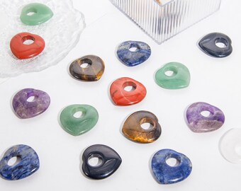 Op de afbeelding: Assortiment van hartvormige kristallen in verschillende kleuren, waaronder groen, rood, paars, bruin en blauw. De stenen hebben een gat in het midden en zijn gerangschikt op een wit oppervlak.