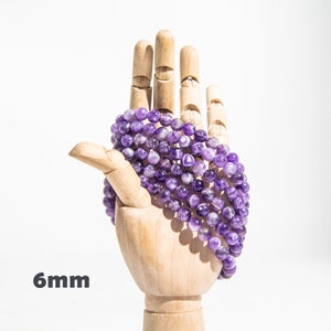 May include: A strand of amethyst beads, 6mm in size, wrapped around a wooden hand. The beads are a deep purple color with white streaks.