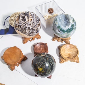 Könnte beinhalten: Eine Sammlung dekorativer Kristallkugeln in verschiedenen Farben und Mustern, präsentiert auf Holzständern. Die Kugeln umfassen eine Geode, einen Moosachat und eine dunkelgrüne Kugel. Ein durchsichtiger Kunststoffbehälter und ein gewebter Untersetzer sind ebenfalls zu sehen.
