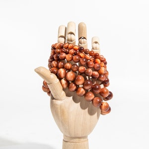 May include: A wooden hand model holding a strand of red tiger's eye beads. The beads are round and have a shiny, metallic finish.