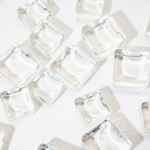 Pode incluir: Uma coleção de azulejos quadrados de vidro transparente, espalhados sobre uma superfície branca. Os azulejos são lisos e reflexivos, com um leve aspecto fosco.
