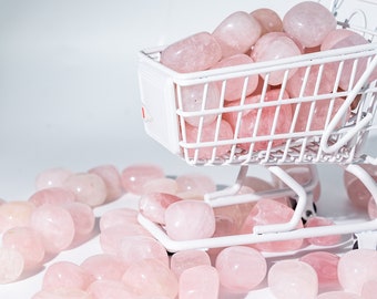 Op de afbeelding: Een miniatuur winkelwagentje gevuld met roze rozenkwarts stenen. De winkelwagen staat op een wit oppervlak met meer rozenkwarts stenen eromheen verspreid.