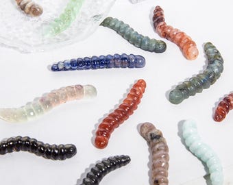 May include: A collection of carved gemstone worms in various colors, including red, blue, green, and brown. The worms are arranged on a white surface, showcasing their detailed, segmented bodies. The gemstones have a polished, translucent appearance.