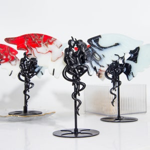 May include: Three black metal sculptures of a couple embracing with wings. The wings are made of red, white, and black marbled resin. The sculptures are on black stands.