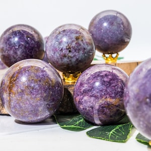 Op de afbeelding: Een verzameling van zes paarse en gouden edelsteenbollen op een houten oppervlak met groene bladeren. De bollen zijn gepolijst en hebben een glinsterende afwerking.