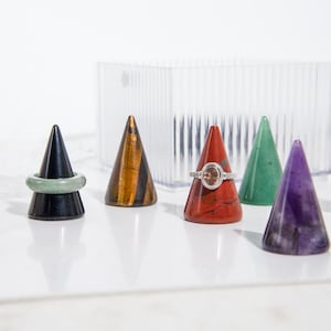 以下が含まれることがあります： さまざまな色と質感の宝石リングホルダーのコレクション。 ホルダーは円錐形で、黒、タイガーアイ、赤、緑、紫の外観をしています。 緑色のリングとダイヤモンドリングがホルダーに表示されています。