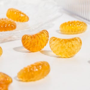Puede incluir: Varias golosinas de goma transl&uacute;cidas de color naranja dorado con forma de gajos de c&iacute;tricos. Cada pieza presenta una superficie texturizada y segmentada que imita la apariencia de un gajo de naranja real, esparcidas sobre una superficie blanca.