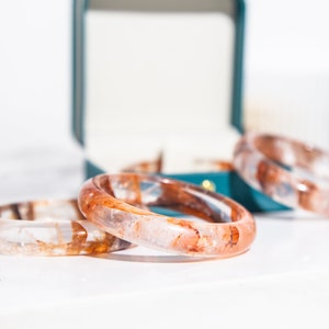 Puede incluir: Tres pulseras de resina transparente con vetas marrones y blancas. Las pulseras están apiladas una encima de la otra.