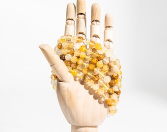 May include: Several bracelets made of yellow and brown gemstone beads are displayed on a wooden hand. The beads vary in color, from light yellow to a deep amber. The bracelets are strung together, creating a layered effect.