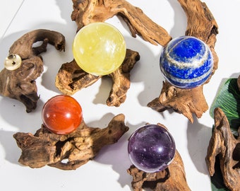 Può includere: Cinque sfere di pietre preziose lucidate esposte su basi in legno naturale. Le sfere sono di colore giallo, rosso, blu, viola e verde chiaro. Le basi in legno hanno un aspetto grezzo e naturale.