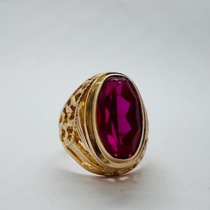 May include: A gold ring with an oval, deep red gemstone. The ring has a textured, ornate band and a polished gold setting. The gemstone is faceted, catching the light and displaying a vibrant colour.
