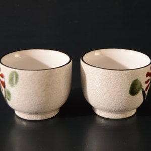 Puede incluir: Dos tazas de t&eacute; de cer&aacute;mica blanca con una superficie texturizada y dise&ntilde;os florales en marr&oacute;n, verde y rojo. Las tazas tienen un borde negro.