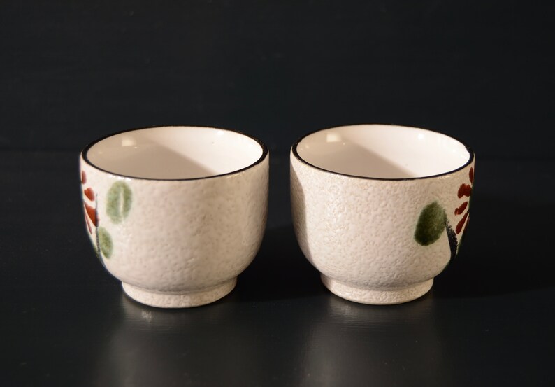 Puede incluir: Dos tazas de t&eacute; de cer&aacute;mica blanca con una superficie texturizada y dise&ntilde;os florales en marr&oacute;n, verde y rojo. Las tazas tienen un borde negro.
