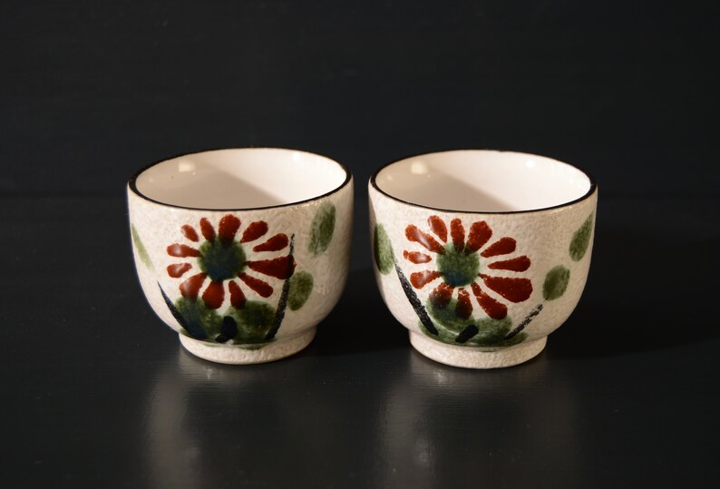 Puede incluir: Dos peque&ntilde;as tazas de t&eacute; de cer&aacute;mica blanca con un dise&ntilde;o floral. Las tazas tienen un patr&oacute;n floral marr&oacute;n, rojo y verde con un contorno negro.