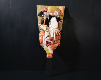 Puede incluir: Un abanico japonés tradicional pintado a mano con una colorida representación de una mujer con un kimono. El abanico está decorado con pan de oro y tiene un mango negro.