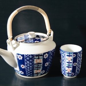 May include: A white teapot with a blue and white floral pattern and a woven handle. A matching blue and white teacup is next to the teapot.