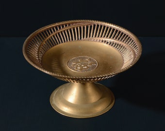 May include: A gold-colored metal bowl with a decorative, openwork design. The bowl sits on a pedestal base.