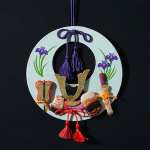 May include: A light blue circular ornament with a gold and purple butterfly, a gold horseshoe, and two purple tassels. The ornament is decorated with purple and green iris flowers and has a gold and red tassel hanging from the bottom.