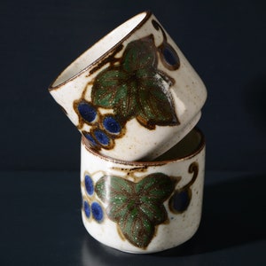 May include: Two white ceramic cups with a brown, green, and blue grapevine design. The cups are stacked on top of each other.