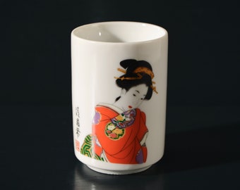 Puede incluir: Taza de cerámica blanca con una ilustración colorida de una mujer con un kimono rojo y morado. La mujer tiene el pelo negro y mira hacia abajo. La taza tiene texto japonés en el lateral.