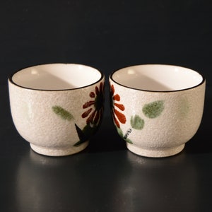 Puede incluir: Dos tazas de t&eacute; de cer&aacute;mica blanca con un dise&ntilde;o floral. Las tazas tienen una superficie texturizada y un borde negro. Las flores son rojas, marrones y verdes.