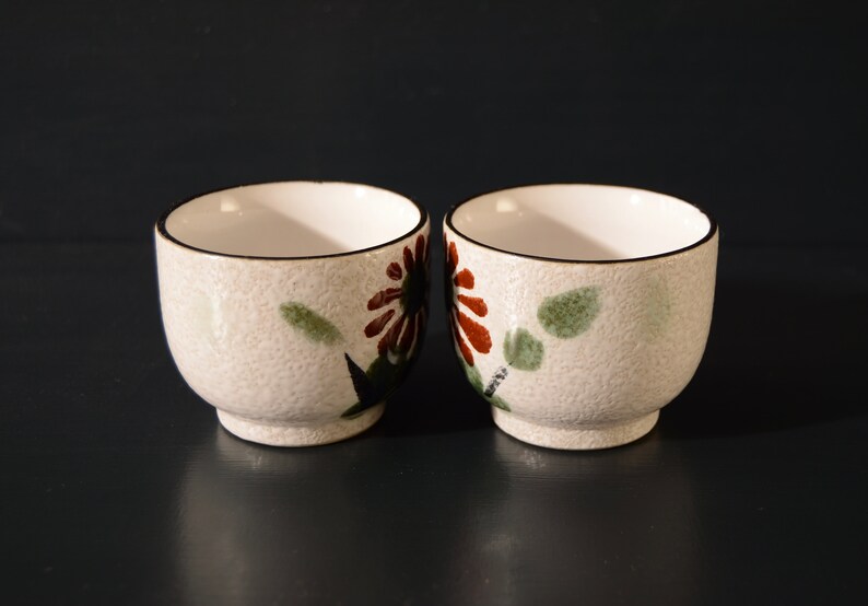 Puede incluir: Dos tazas de t&eacute; de cer&aacute;mica blanca con un dise&ntilde;o floral. Las tazas tienen una superficie texturizada y un borde negro. Las flores son rojas, marrones y verdes.