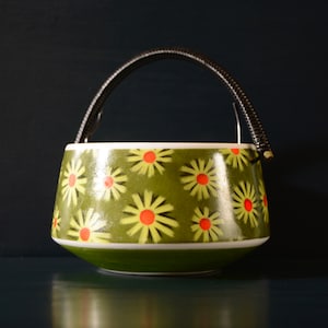 May include: A green ceramic sugar bowl with a black handle and a daisy pattern. The daisies have yellow petals and red centers.