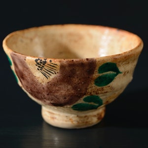 May include: A small, brown and white ceramic bowl with green and black painted designs. The bowl has a rounded shape and a slightly flared rim.