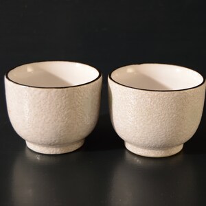 Puede incluir: Dos peque&ntilde;as tazas de cer&aacute;mica blanca con una superficie texturizada y un borde negro. Las tazas est&aacute;n sobre una superficie negra.