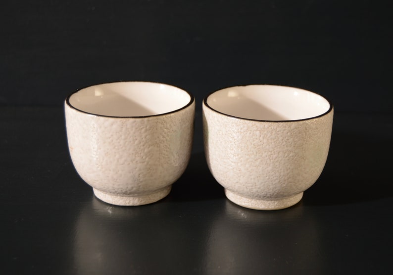 Puede incluir: Dos peque&ntilde;as tazas de cer&aacute;mica blanca con una superficie texturizada y un borde negro. Las tazas est&aacute;n sobre una superficie negra.