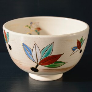 May include: A white ceramic bowl with a decorative design of colorful leaves. The bowl has a slightly rounded shape and a small foot.